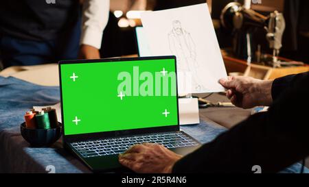 Craftsman using greenscreen on laptop, experienced tailor looking at chroma key display in workshop. Couturier designing items with isolated mockup template in tailoring atelier. Stock Photo