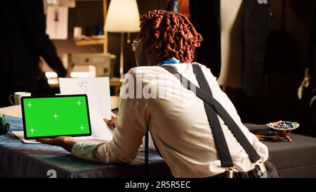 Young tailor checking greenscreen in atelier workshop, using isolated chroma key display at workstation table. Professional designer working with mockup template on tablet, handcraft. Stock Photo
