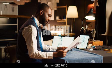 Couturier comparing sketches with tablet, looking at drawings of clothes in tailor atelier. Fashion designer crafting clothing line and items using textile industry tools. Handheld shot. Stock Photo