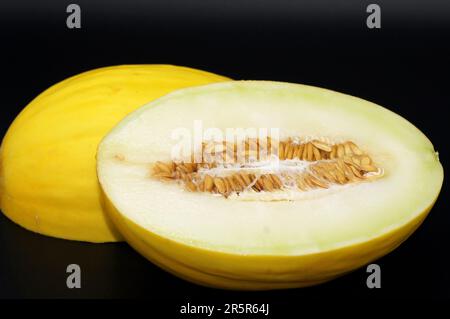 Two ripe yellow melons cut in half, revealing juicy red interiors, on a black surface with a bite taken out of one of the melons Stock Photo