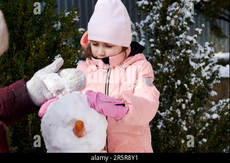 Cute little child girl in warm pink down jacket, enjoying making a snowman in a backyard, spending happy winter holidays Stock Photo
