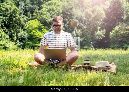 Young adult man freelancer wearing sunglasses working on the laptop while sitting on the lawn in the green park in summer. Stock Photo