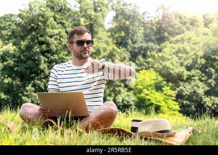 Time saving and planning, man working on a laptop and looking at his wristwatch while sitting on the lawn working outdoors in the park. Stock Photo