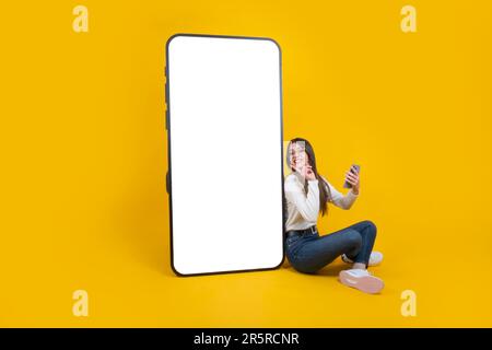 cheerful woman leaning big smartphone with empty blank screen. Huge mobile phone mockup. Lady sitting on floor holding phone in hand, pointing display Stock Photo