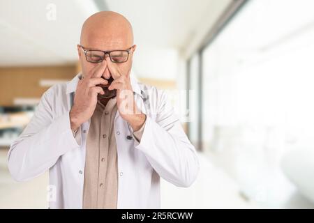 Caucasian bald middle aged frustrated doctor in his room. Tired, stressed, bored senior physician rubbing his eyes. Blurred background, copy space. Stock Photo