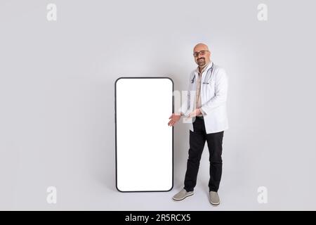 Male doctor presenting new hospital advertising concept idea image. Full length body shot of physician showing big smartphone empty screen, mock up. Stock Photo