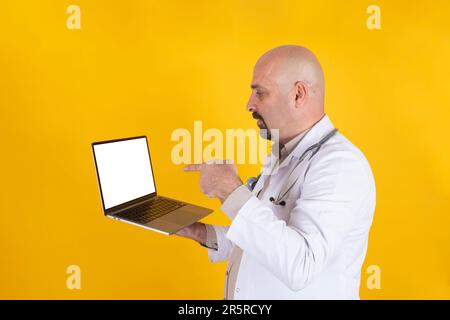 Middle aged bald doctor pointing laptop screen. Caucasian male physician holding modern notebook with empty blank white screen mock up. Stock Photo