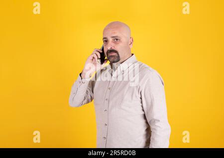 Standing over yellow studio background. Bald male seriously listening call. Businessman talking with client. Copy space. Stock Photo