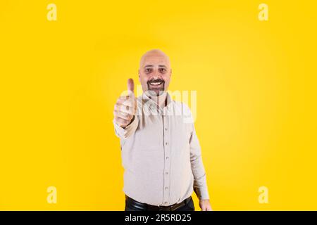 Middle aged bald man doing happy thumbs up gesture approving expression. Wearing casual beige shirt isolated over yellow background, copy space. Stock Photo