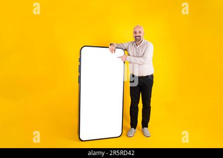 Full body length body shot man leaning big smartphone, touching white empty screen display panel with finger. Looking camera. Mobile phone mock up. Stock Photo