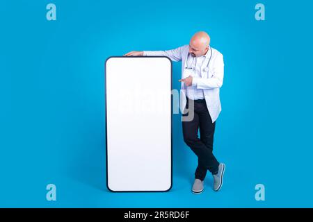 Middle aged male doctor, leaning and pointing big huge mobile phone with blank screen. Full size physician standing over isolated blue background. Stock Photo