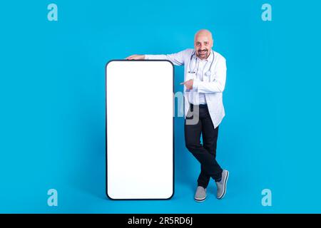 Cheerful smiling middle aged doctor pointing big smartphone. Bald physician leaning huge mobile phone with empty screen, mock up. Stock Photo