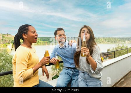 Group of happy young multiracial friends colleagues freelancers in casual clothes drinking cold beer and dancing together while having party on buildi Stock Photo