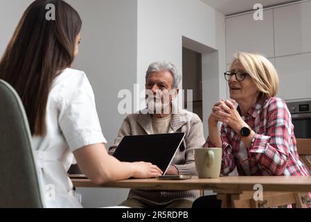 Female professional doctor consulting senior couple patient during medical care visit. Young woman physician talking providing medical assistance sitt Stock Photo