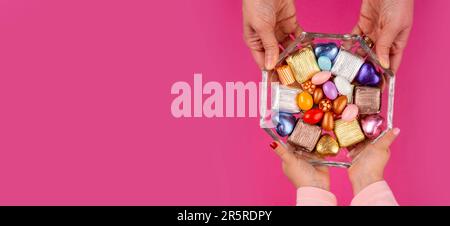 Mother and daughter hands holding bowl of candies. Top view, isolated pink background. Copy space. Many types wrapped luxury chocolate, almond sweet. Stock Photo