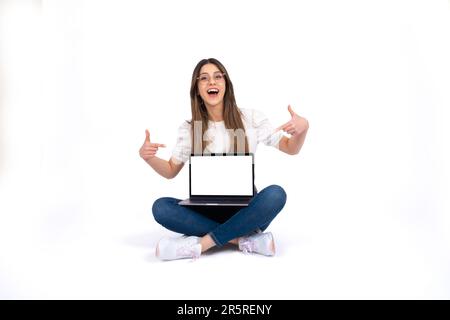 Teen girl sitting on the ground and showing laptop. Cross-legged posing on isolated white background. Fingers pointing empty screen notebook mock up. Stock Photo