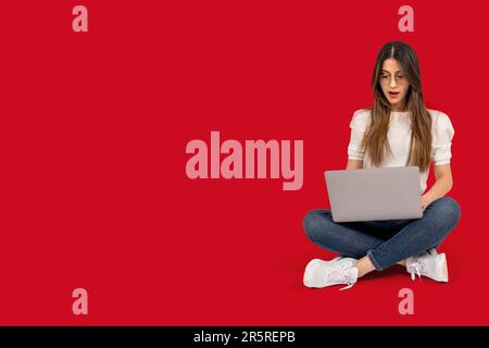 Full body length view of shocked woman. Brunette young girl sitting on the ground. Holding laptop looking screen surprised what she see. Stock Photo