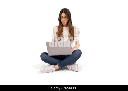 Shocked young woman, full body shot of shocked young woman. Sitting on the floor, using laptop. Lifestyle concept. Holding notebook. Stock Photo