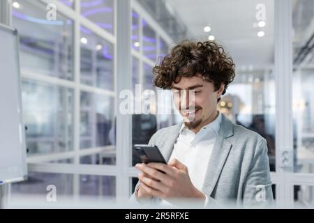Successful and happy businessman with beard works in modern bright office, male boss uses laptop, business owner in casual shirt holds phone, browses internet pages. Stock Photo