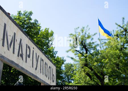 The name of the city in Ukrainian Mariupol on a road sign broken from bullets against the background of the flag of Ukraine in the sky, an occupied ci Stock Photo