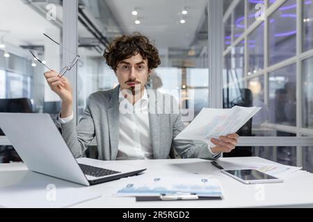 Dissatisfied and angry businessman inside office showing reports and bills documents to camera, boss in shirt looking at camera working with laptop paper work Stock Photo