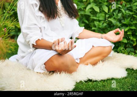 Middle age woman meditating in green garden, sitting on sheep skin Stock Photo