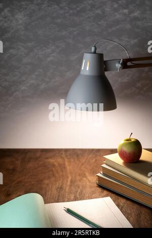 table lamp illuminates the workplace on the office desk, a stack of books, an open notebook, a pencil and an apple on a gray background with clouds Stock Photo