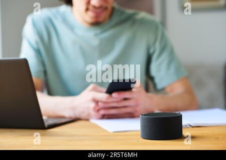 Close-up of young man connecting smart speaker to his smartphone while sitting at table with laptop Stock Photo