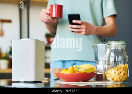 Close-up of young man connecting smart speaker to phone to listen to music while standing in the kitchen Stock Photo