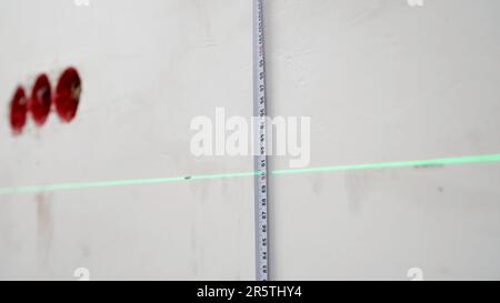 A man uses a cross laser level and a tape for accurate measurements on a light wall, close-up. Measure the height with a laser level. Laser level and Stock Photo