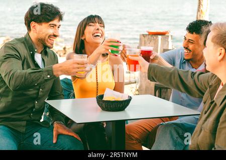 A diverse group of friends sitting in a beachfront bar, cheerfully toasting with vibrant cocktails in plastic cups. The lively scene showcases the joy Stock Photo