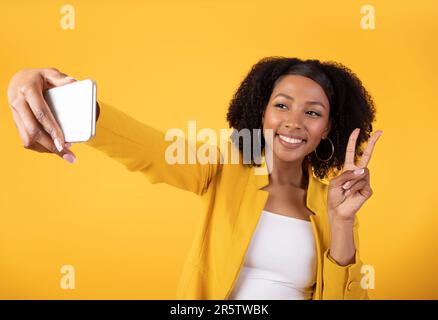 Cheerful young black lady taking selfie on cellphone, showing peace gesture and smiling, updating social media profile Stock Photo
