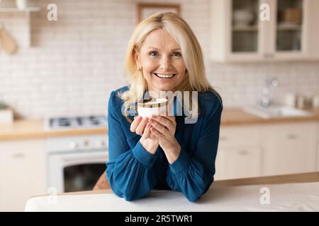 Cheerful mature woman enjoying herbal tea at home, standing at cozy kitchen, holding coffee mug and smiling at camera Stock Photo