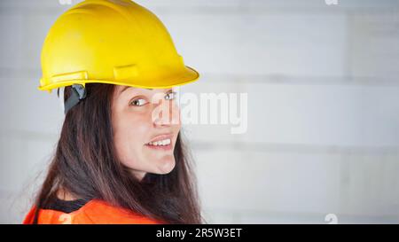 Young woman in yellow hard hat and orange high visibility vest, long dark hair, looking over her shoulder, smiling confident. Blurred construction sit Stock Photo