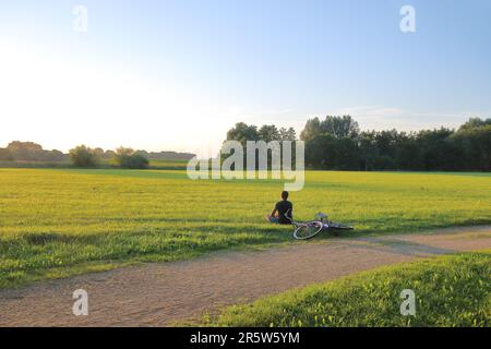 In the photo seen a young man sitting on the meadow and admiring the sunset. Near visible bicycle lying on the grass. Stock Photo