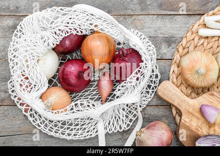 Wooden board and string bag with different kinds of onion on white tile ...