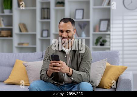 Young smiling African American man sitting on sofa at home and using phone, typing messages, texting, playing online games. Stock Photo