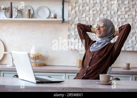 Rest after distance learning. Young Muslim female student in hijab sits at home at the table in front of the laptop and relaxes. She threw her hands behind her head, closed her eyes, a break. Stock Photo