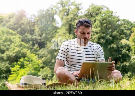 Young adult student sitting on lawn in the park and watching online lesson on the internet using his laptop. Stock Photo
