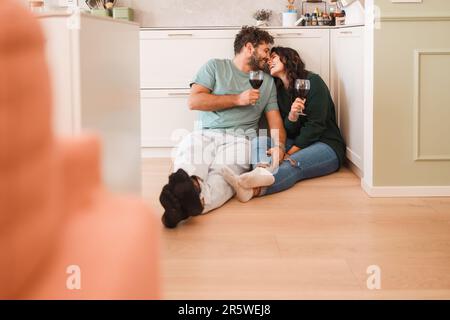 Happy Newlyweds in Bright Kitchen - Joyful newlywed couple sitting on the floor of their kitchen. They share a tender moment as they playfully touch n Stock Photo