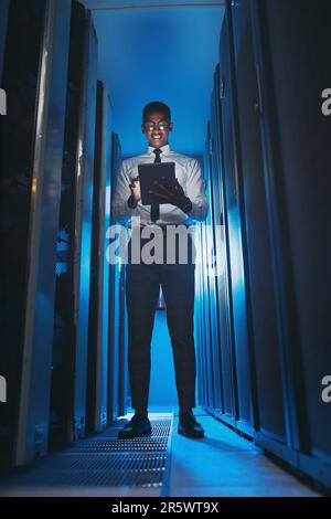 The server room is my home. Full length shot of a young IT specialist standing alone in the server room and using a digital tablet. Stock Photo