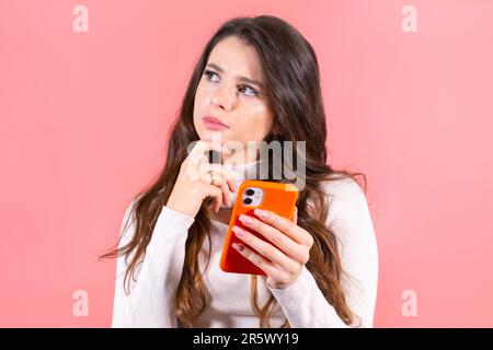 Young woman with amused and smiling expression enjoys scrolling social media on orange smartphone. Long-haired brunette lady smiles looking at feeds Stock Photo