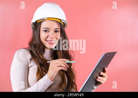 Brown-haired woman in white hardhat with smile prepares project for construction. Young lady engineer writes information on black clipboard Stock Photo