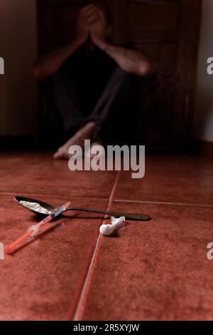 Man lying on the floor in the dark with his hands on his head desperate for drugs in the foreground Syringe with heroin and spoon Stock Photo