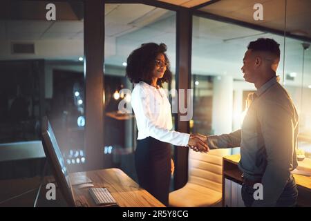 Thank you for helping me handle those deadlines. two businesspeople shaking hands in an office at night. Stock Photo