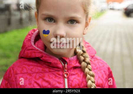 Little girl with drawing of Ukrainian flag on face in heart shape outdoors Stock Photo