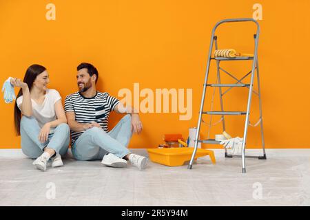 Happy designers sitting on floor with painting equipment near freshly painted orange wall indoors Stock Photo