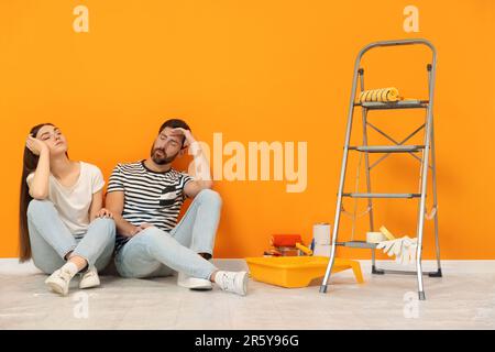 Tired designers sitting on floor with painting equipment near freshly painted orange wall indoors Stock Photo