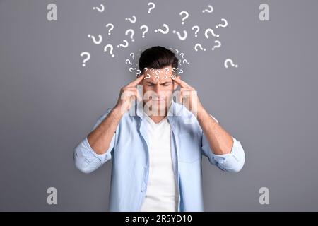 Amnesia concept. Man surrounded by question marks trying to remember something on grey background Stock Photo