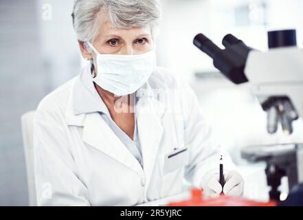 This is my work uniform. Portrait of a focused elderly female scientist making notes while looking into the camera and wearing a doctors mask inside Stock Photo
